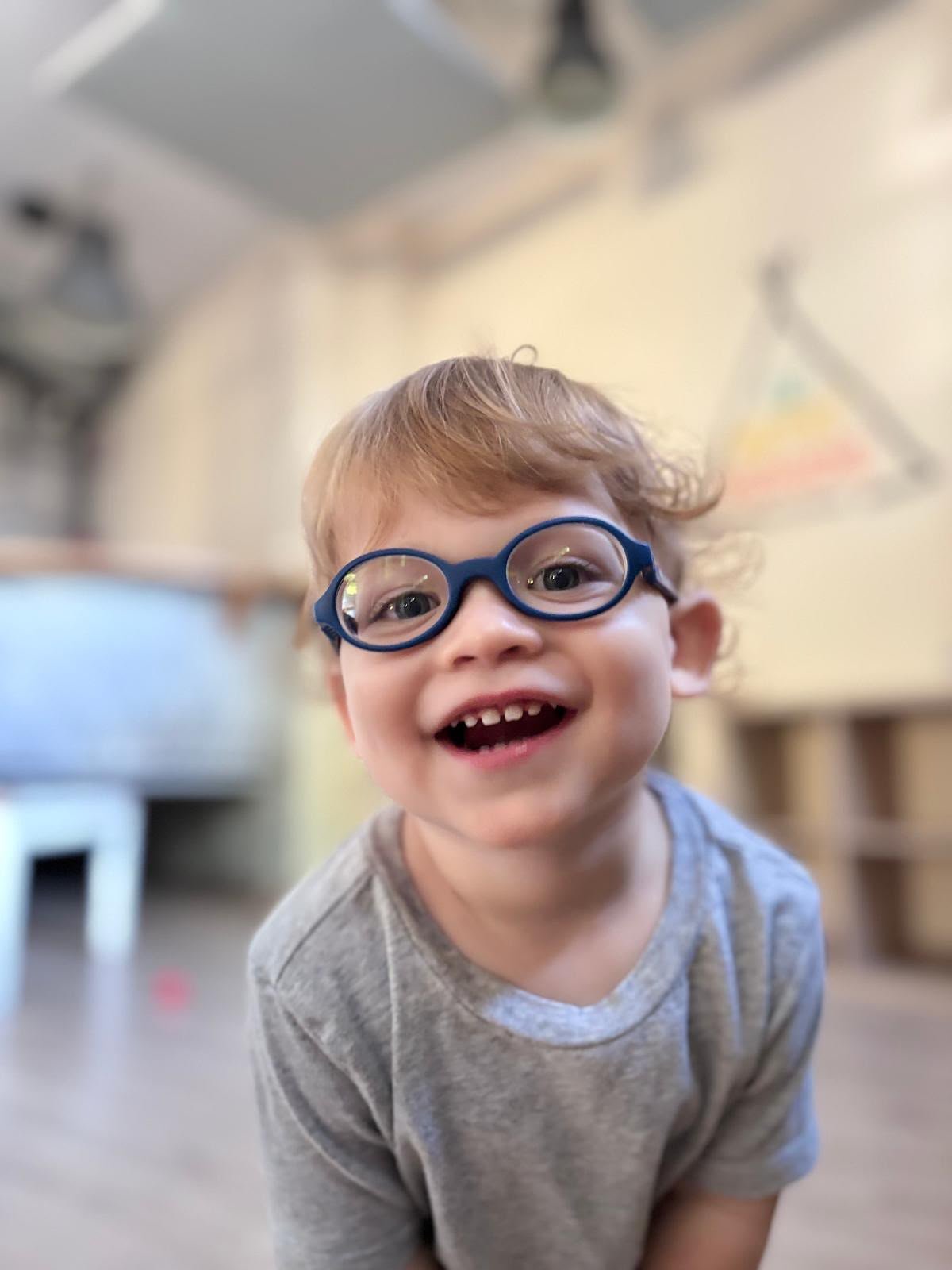Niño pequeño con gafas azules sonriendo a la cámara en una sala de Le Petit Monde